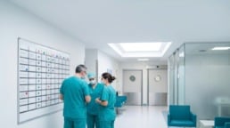 Three clinicians in scrubs huddle beside a schedule board in an ambulatory surgery center hallway