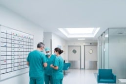 Three clinicians in scrubs huddle beside a schedule board in an ambulatory surgery center hallway